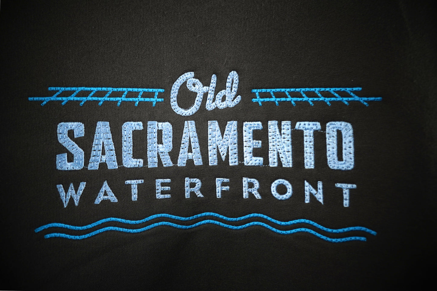 The words "Old Sacramento Waterfront" embroidered in light blue on a dark gray background. There are two train track segments embroidered with teal blue thread on either side of the word old and two wavy teal blue lines at the bottom of the design to represent water. Individual stitching detail can be seen in each element.