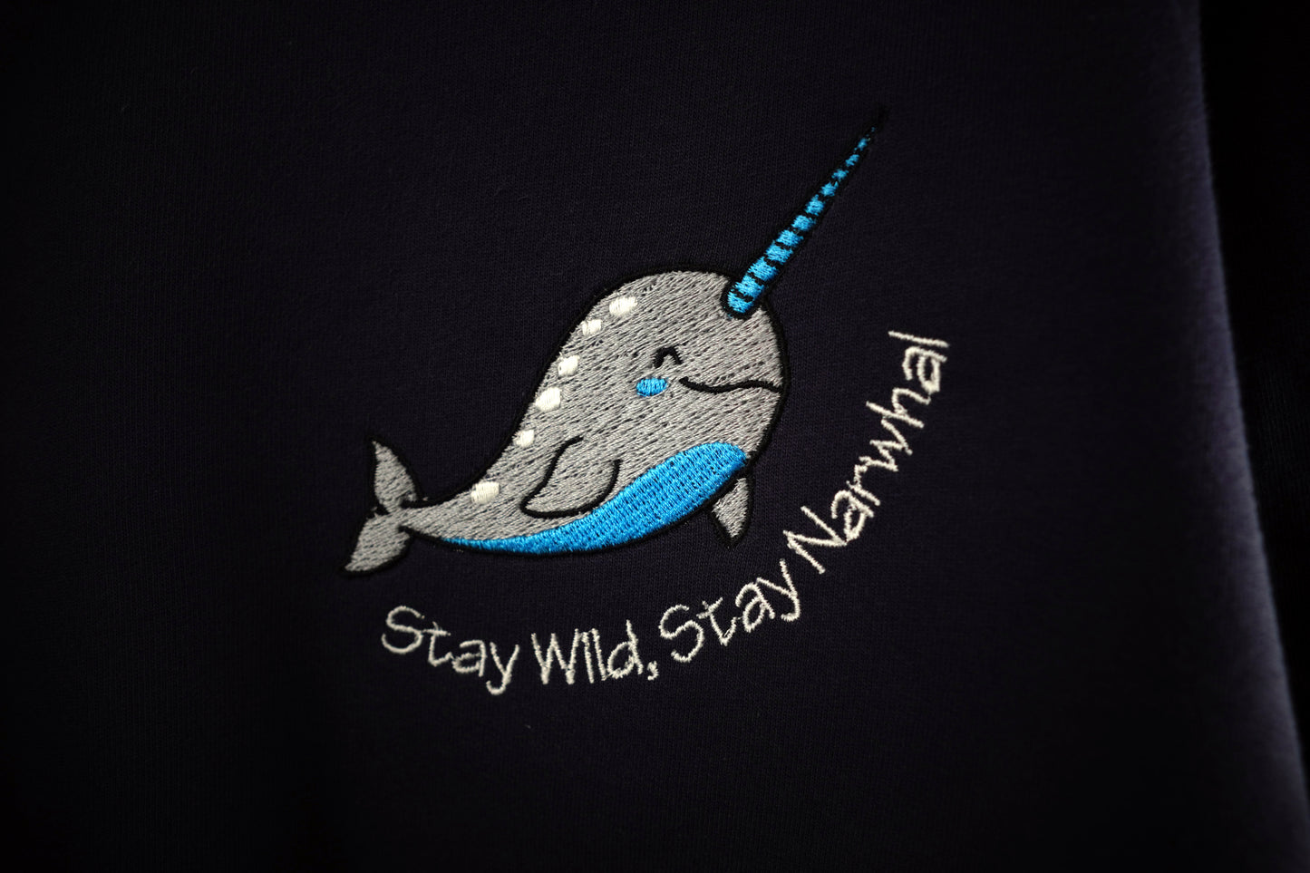 Grey and teal embroidered Narwhal design on a navy blue background with the words Stay Wild, Stay Narwhal in white lettering under the Narwhal. The photograph is close enough to see the stitching detail from the embroidery.