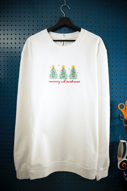 White crewneck sweatshirt with a Christmas tree design embroidered in the center chest and Merry Christmas in a handwritten-style script embroidered in red below the trees. The sweatshirt is hanging on a wooden hanger in front of a dark aquamarine pegboard background.