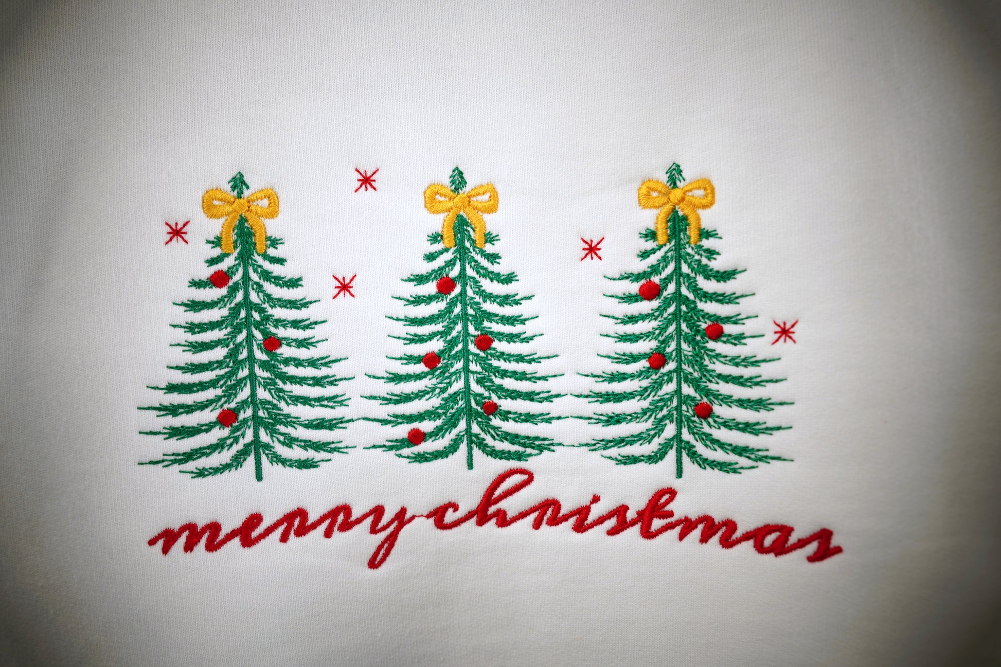 Three evergreen trees embroidered in green thread on a white crewneck sweatshirt. Each segment of the branches is visible in the photo. The trees are topped with a golden bow and accented with tiny red ornaments. Red stitched stars dot the sky. Merry Christmas in a handwritten-style script is stitched in red below the trees.