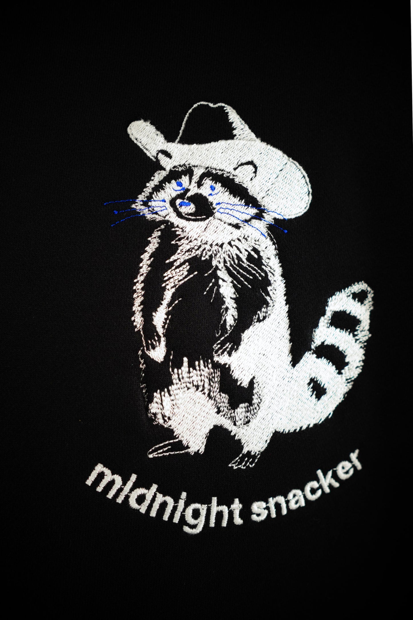 Closeup photo of an artistic line drawing rendition of a raccoon weathering a cowboy hat with the words "midnight snacker" under the raccoon. The design was embroidered onto a black hoodie using white thread. The raccoons whiskers and eyes have been stitched with blue thread. The stitching detail is clearly visible throughout the image.