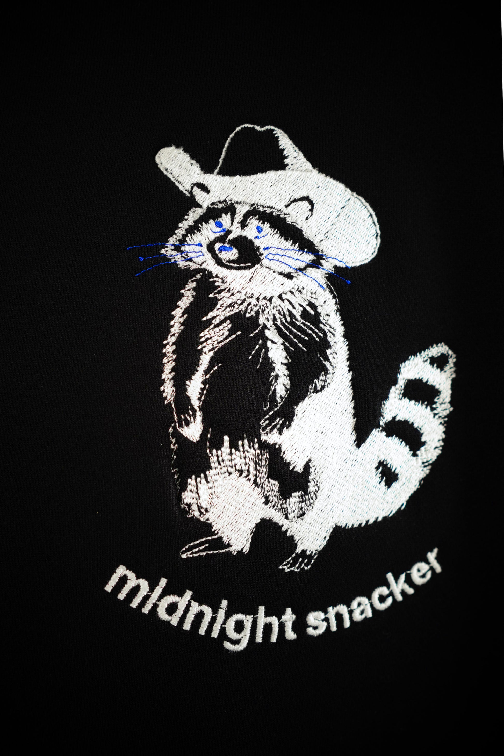 Closeup photo of an artistic line drawing rendition of a raccoon weathering a cowboy hat with the words "midnight snacker" under the raccoon. The design was embroidered onto a black hoodie using white thread. The raccoons whiskers and eyes have been stitched with blue thread. The stitching detail is clearly visible throughout the image.