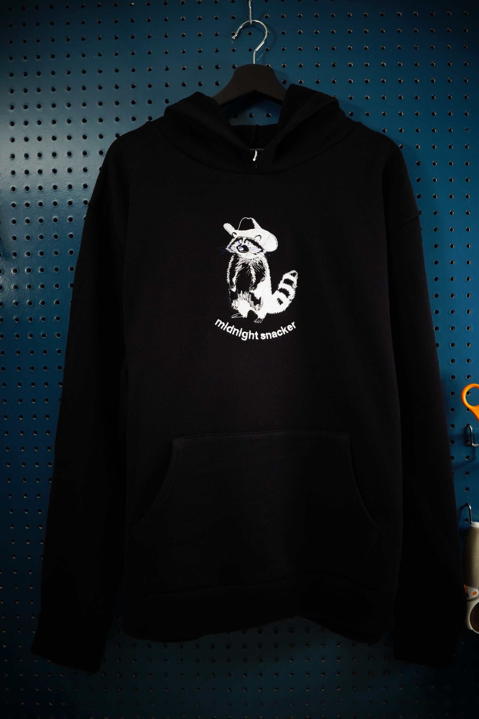 Artistic line drawing rendition of a raccoon weathering a cowboy hat embroidered onto a black hoodie using white thread with the words "midnight snacker" under the raccoon. The hoodie is hanging on a wooden hanger in front of a dark aquamarine pegboard wall. 