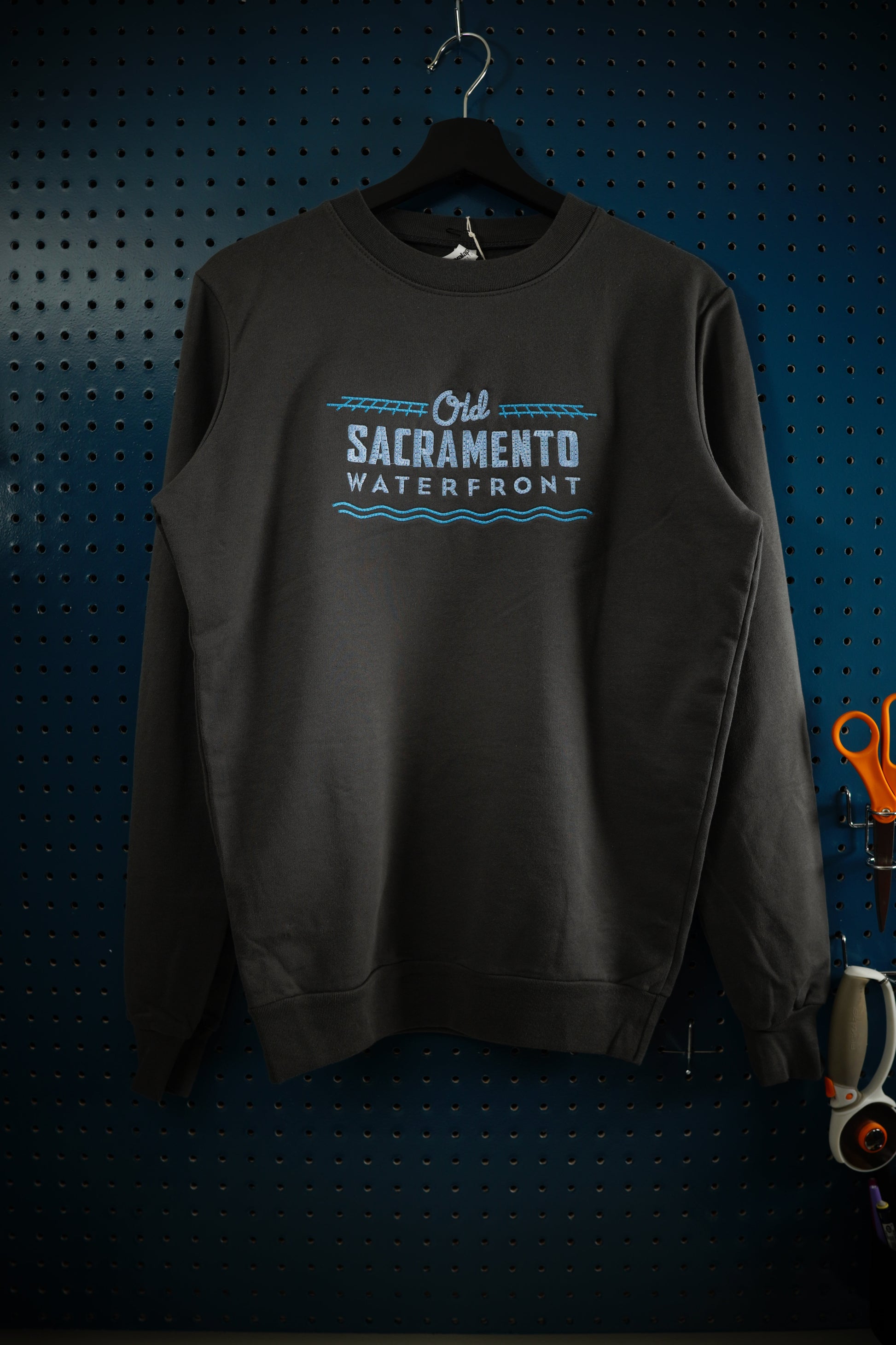 Old Sacramento Waterfront two-tone blue design embroidered onto a dark gray crewneck sweatshirt. The sweatshirt is hanging on a wooden hanger in front of a dark aquamarine pegboard.