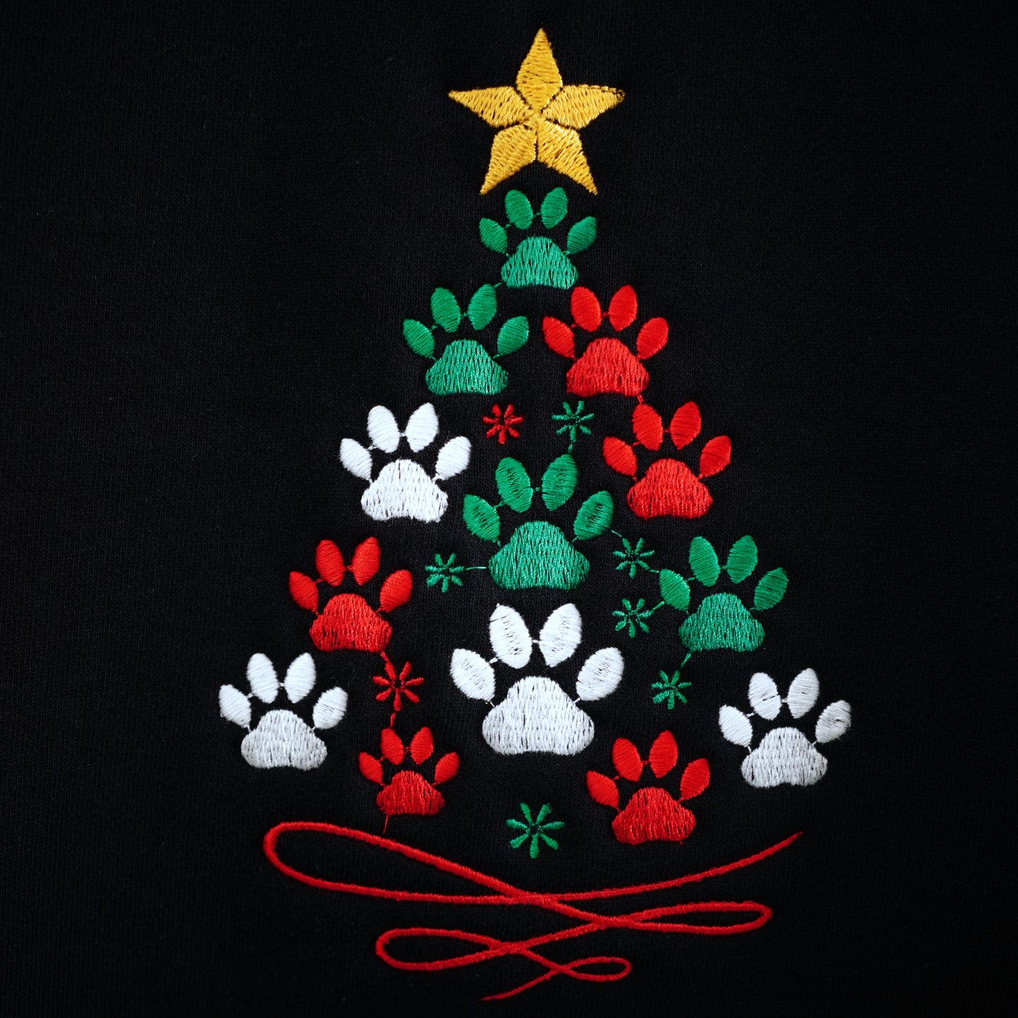 Closeup photo of embroidered Christmas tree design made of green, red and white paw prints with a yellow star on top on a black background. The stitch patterns formed by the embroidery is clearly visible showing the detail of the design.