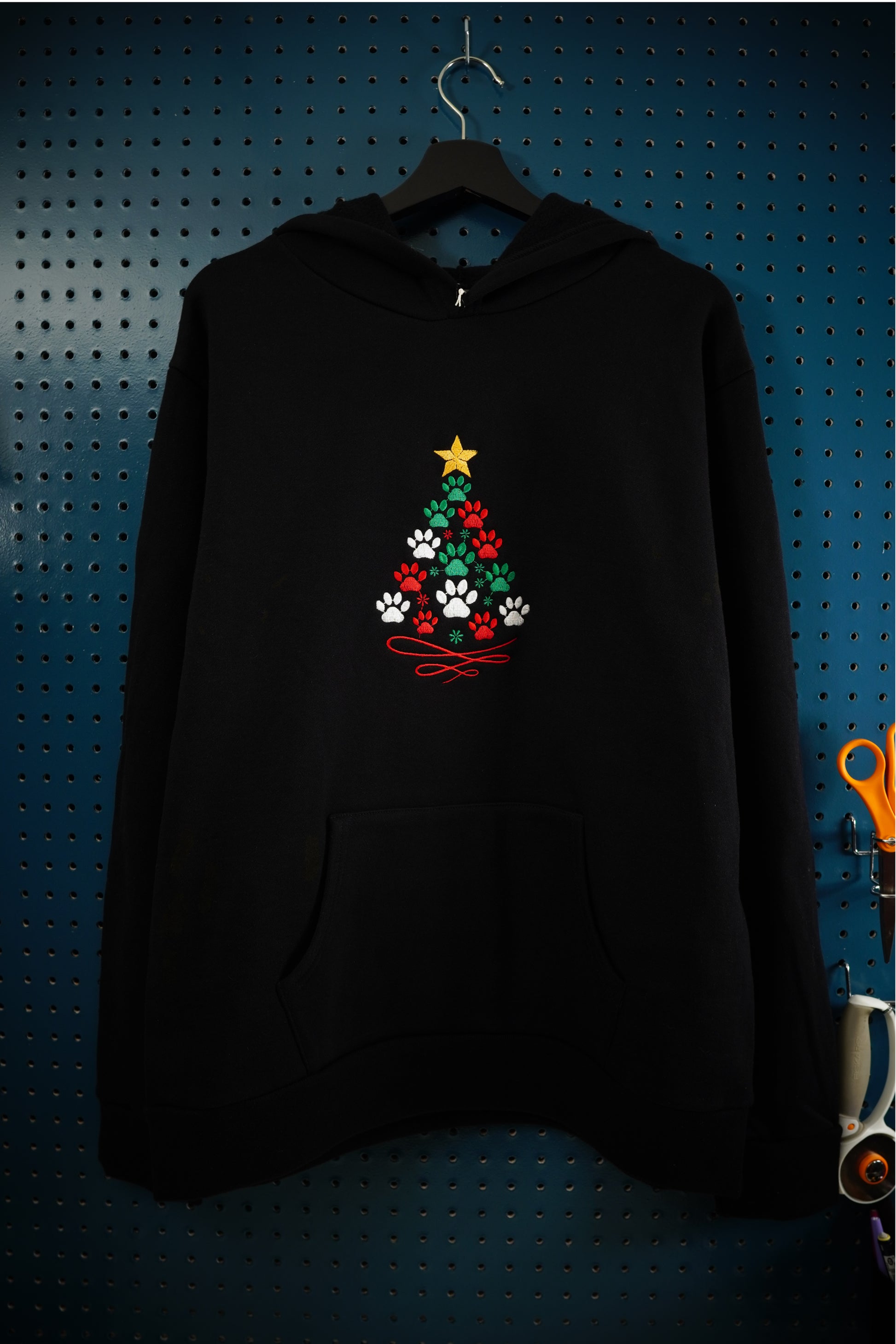 Black hoodie with festive holiday tree embroidered design formed by green, red and white paw prints with a gold star on top, hanging on a wooden hanger against a dark aquamarine pegboard background.