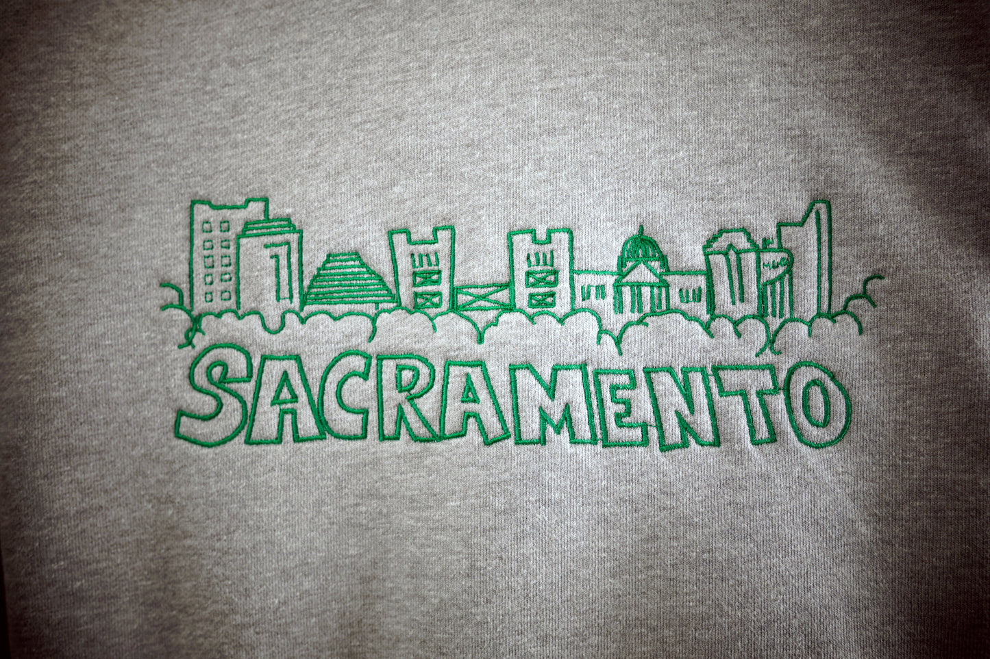 Closeup photo of a hand drawn artistic rendition of the Sacramento skyline embroidered onto a heather gray hoodie using green thread, with the word Sacramento below the drawing. The stitching detail can be seen in the photograph. 