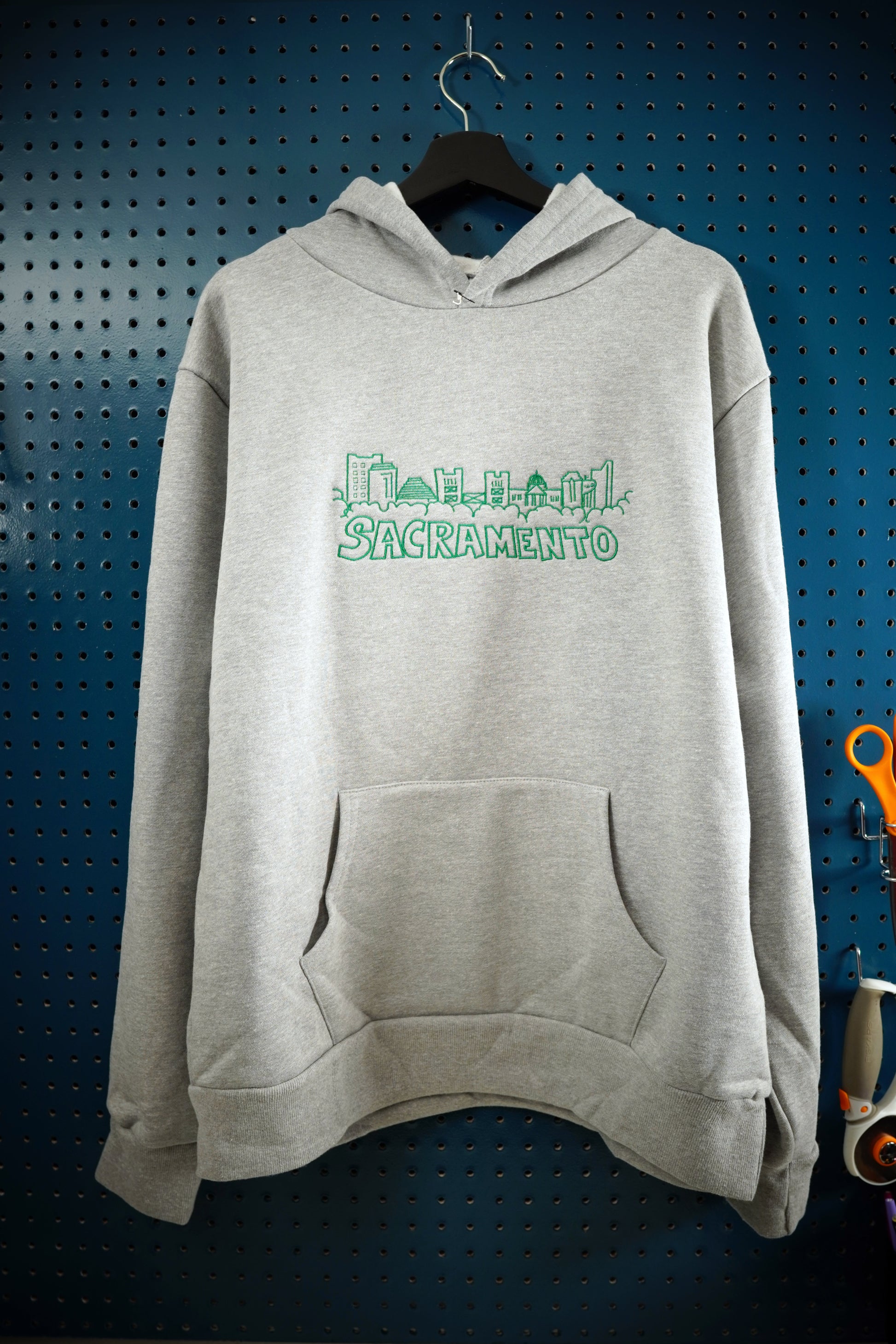 Hand drawn artistic rendition of Sacramento skyline embroidered onto heather gray hoodie using green thread, with the word Sacramento below the drawing. The hoodie is hanging on a wooden hanger against a dark aquamarine pegboard wall. 