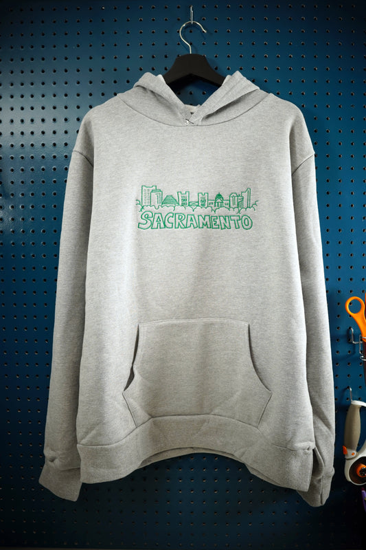 Hand drawn artistic rendition of Sacramento skyline embroidered onto heather gray hoodie using green thread, with the word Sacramento below the drawing. The hoodie is hanging on a wooden hanger against a dark aquamarine pegboard wall. 