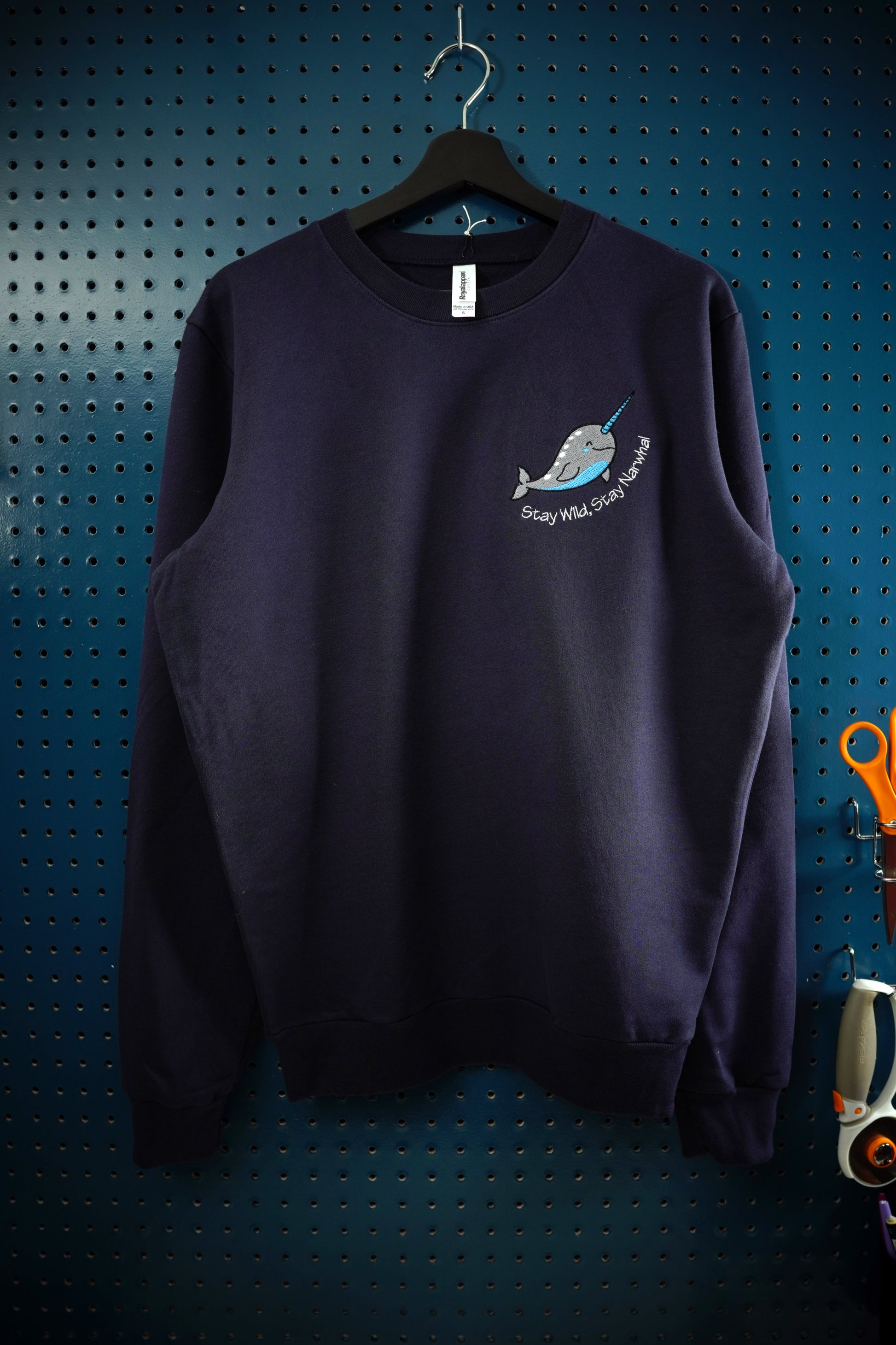 Teal and gray Narwhal logo embroidered in the left chest position on a navy blue crewneck sweatshirt. The phrase Stay Wild, Stay Narwhal is embroidered under the whale in white lettering. The sweatshirt is hanging on a wooden hanger in front of a dark aquamarine pegboard.