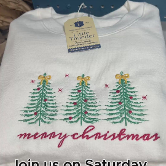 Timelapse video of three evergreen trees being embroidered onto a white crewneck sweatshirt with Merry Christmas in a handwritten-style script embroidered in red below the trees. 